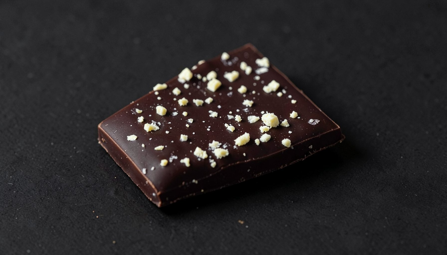Dark chocolate bar with pale cream flecks on a charcoal background, bold and satisfying